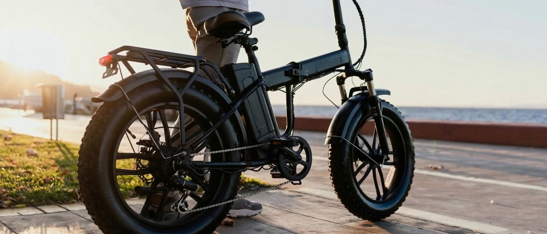 A rider on a black folding e-bike cruising along a seaside promenade at sunset
